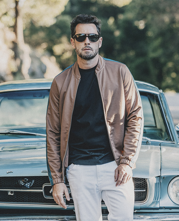 Cool man with brown jacket in bomber style and brown sunglasses in retro design, leaning on a old mustang car